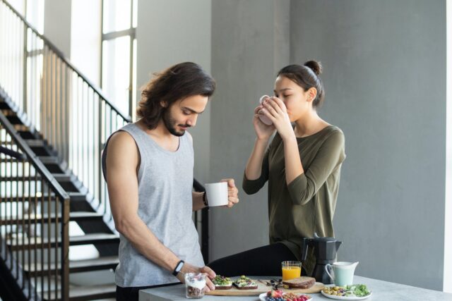 Couple enjoying breakfast with coffee – man in a grey tank top and woman in a green shirt at a table with plates of food